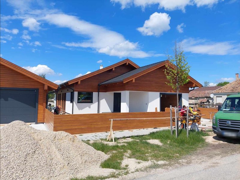 Das fertige Haus von der Straße: holzverkleidete Giebel, überdachter Eingang und Garage; der Zaun entsteht.