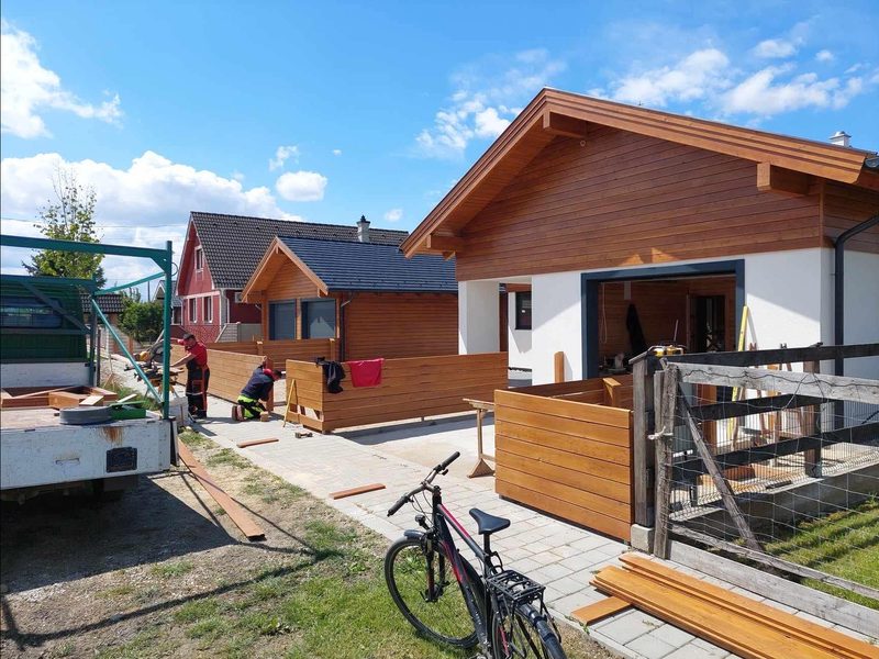 Die offene Garage des Hauses, dahinter das holzverkleidete Nebengebäude, vorn der entstehende Holzzaun.
